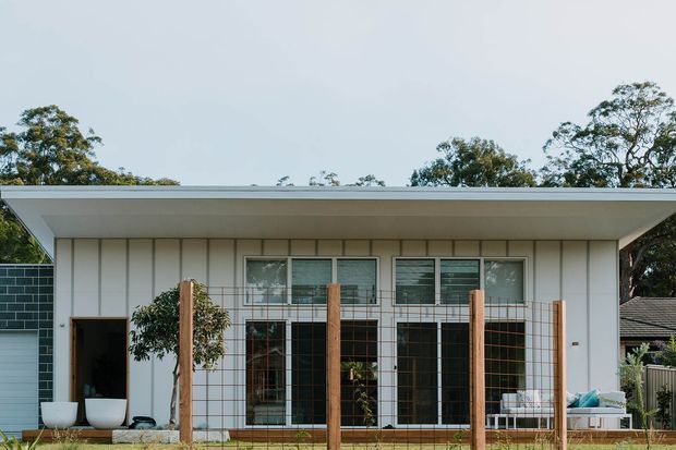 Single-storey contemporary residence viewed from the front, with a broad low-pitched or flat roof, white vertical panel cladding, large dark-framed glazed openings, and a timber-post wire fence. A covered outdoor seating area and landscaping sit in front of the house.
