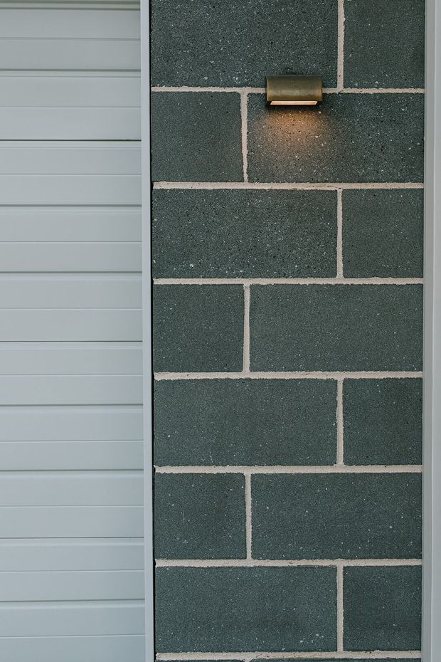 Close-up exterior detail showing dark grey textured brick masonry with light mortar joints, beside pale grey horizontal weatherboard cladding. A small bronze wall light provides a warm glow on the brick surface.
