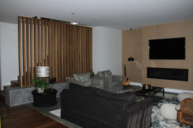 Contemporary living room with a dark sectional sofa, grey armchairs, patterned area rug, and a low black coffee table. A vertical timber-slat divider separates the seating area from the adjacent space, while a timber-panelled media wall incorporates a television and built-in fireplace. The room has a white ceiling, dark timber flooring, recessed lighting, and layered neutral furnishings.