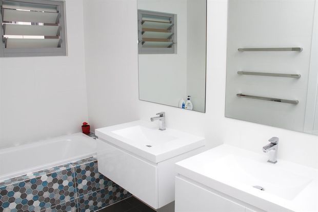 Bright contemporary bathroom featuring a double vanity with white integrated basins, wall-mounted mirrors, chrome tapware and towel rails, a bathtub, and a geometric hexagonal mosaic tile accent around the bath. Louvered windows provide ventilation and natural light.