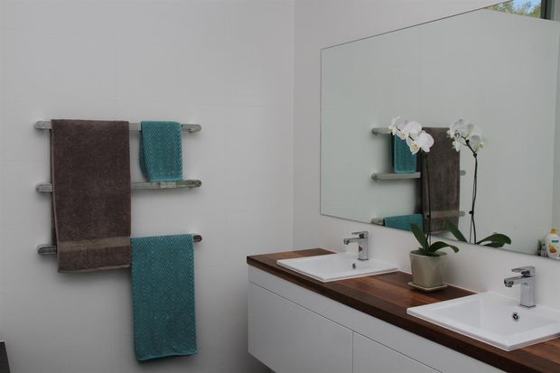 Bright contemporary bathroom with a long white double vanity, two integrated rectangular basins, dark timber benchtop, large wall mirror, chrome taps, and wall-mounted heated towel rails holding brown and teal towels. An orchid adds a small decorative touch.