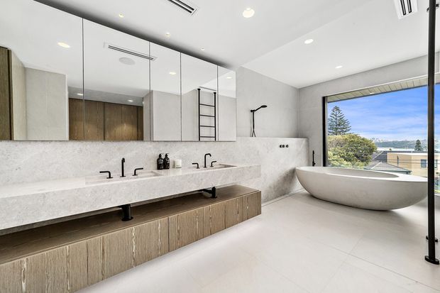Contemporary double-vanity bathroom with a freestanding oval bathtub, expansive mirrored wall, pale stone surfaces, warm wood-effect floating cabinetry, black tapware, and large glazing overlooking a coastal suburban view.