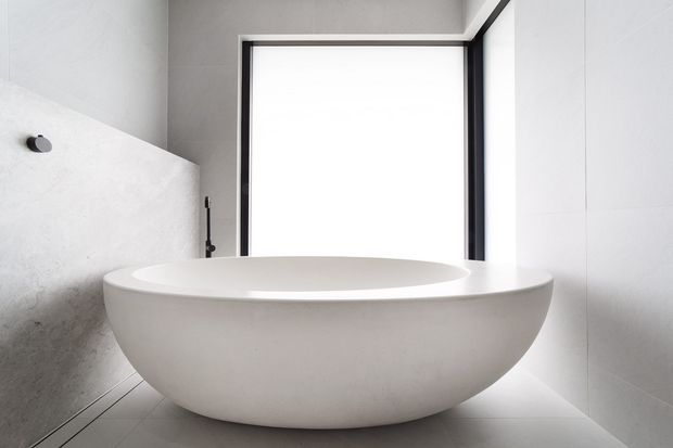 A minimalist contemporary bathroom featuring a large freestanding oval bathtub in a pale smooth finish. Light grey and white wall and floor surfaces surround the tub, with a dark-framed window providing a strong vertical contrast.