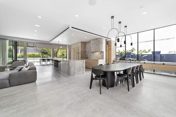 A spacious open-plan contemporary interior combining kitchen, dining, and living zones. The room features a large stone kitchen island, integrated light timber cabinetry, a long dark dining table with black chairs, grey upholstered sofas, expansive floor-to-ceiling glazing, and sculptural pendant lighting. The pale polished concrete floor and abundant daylight create a refined, airy atmosphere.