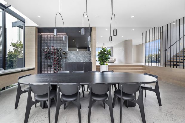 A spacious contemporary dining area with a long dark dining table and sculptural black chairs, illuminated by suspended pendant lights. The room features polished concrete flooring, light timber wall and stair finishes, extensive glazing, a dark feature ceiling, built-in wine storage, and a partially screened staircase leading to an upper level.