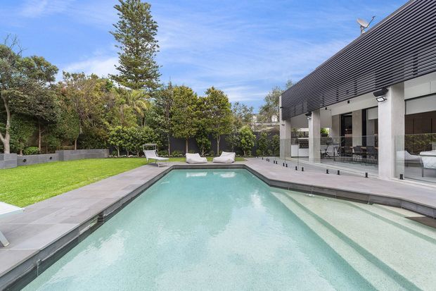 Contemporary residential pool courtyard with a long rectangular swimming pool, pale stone paving, manicured lawn, landscaped screening and a modern house featuring a dark horizontal louvred facade, broad glazing and covered outdoor living areas.