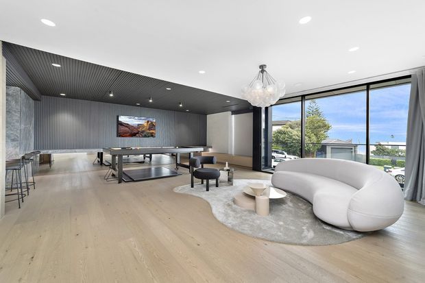 A bright, spacious contemporary open-plan living and recreation area with pale timber flooring, a sculptural white curved sofa, round coffee table, soft grey rug, and a pool table beneath a dark ribbed ceiling and feature wall. Floor-to-ceiling glazing opens to an exterior suburban view, while a clustered glass pendant adds a decorative focal point.