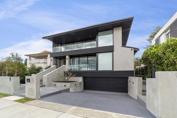 Exterior view of a contemporary multi-level residence with a dramatic asymmetrical overhanging roof, dark horizontal cladding, pale rendered walls, extensive glazing, glass balustrades, and an integrated double garage. A broad concrete driveway and stepped front entry define the street-facing approach.