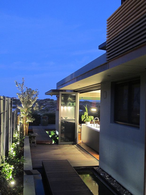 Twilight exterior view of a contemporary house showing a timber deck and narrow water feature leading toward an open, illuminated kitchen pavilion. The building has clean flat roof planes, projecting eaves, dark horizontal timber screening on the upper facade, large glazing, and integrated landscape lighting. Planting and a boundary fence line the walkway, with hills visible in the distance.