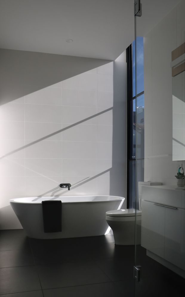 A contemporary bathroom featuring a freestanding white bathtub, wall-mounted dark tapware, toilet, floating white vanity, large mirror, and a frameless glass shower screen. Large pale wall tiles are sharply illuminated by sunlight, contrasting with dark floor tiles. A tall narrow window brings in strong natural light and urban views.