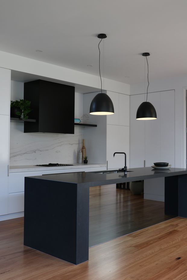 A contemporary minimalist kitchen centered on a large dark island with an integrated sink and black tap. The kitchen features white full-height cabinetry, a pale veined stone splashback and benchtop, warm timber flooring, a black wall-mounted rangehood, and two oversized black pendant lights. Simple styling and clean lines create a refined monochrome interior with warm wood contrast.