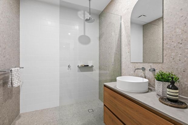 Contemporary bathroom featuring a walk-in shower with clear glass screen, white vertical and small-format wall tiles, terrazzo-like textured wall and floor surfaces, and a timber floating vanity with a white vessel basin. A large frameless mirror, wall-mounted tapware, greenery, and neutral finishes create a clean, refined atmosphere.