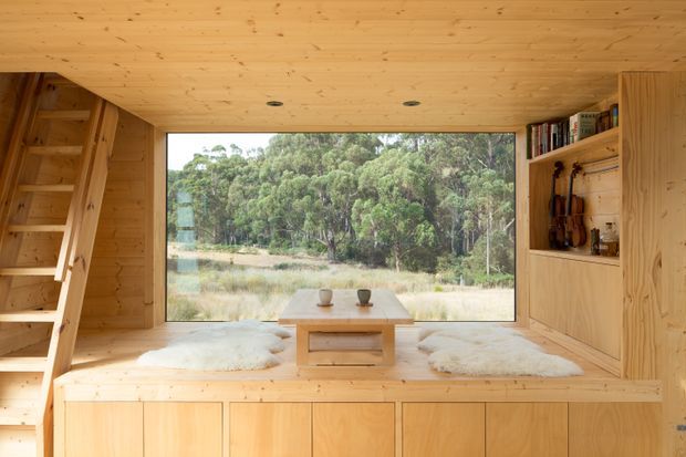 A compact, warm timber-lined interior with a large picture window overlooking a wooded natural landscape. Built-in cabinetry and shelving frame a raised window seat, with a small timber table, stools, and pale sheepskin rugs. A timber ladder at left leads to an upper level or loft, suggesting a cabin-like retreat.