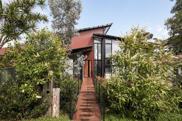 Exterior view of a compact, multi-level contemporary dwelling approached by a narrow reddish timber or metal walkway through dense landscaped vegetation. The building features dark-framed glazing, vertical red corrugated cladding, a sharply sloping asymmetrical roofline, and a sheltered entry porch. Mature trees and shrubs partially screen the facade.