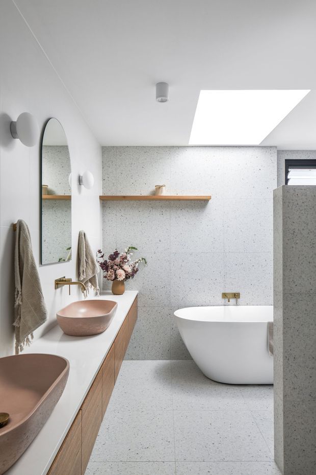 Bright contemporary bathroom with a freestanding white bathtub, double vessel basins on a timber vanity, a large arched mirror, brass fixtures, pale terrazzo-style wall and floor surfaces, and a large rectangular skylight. A partial shower enclosure is visible at right.