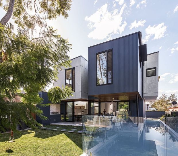 Exterior view of a contemporary multi-level residence with a strongly articulated dark vertical-clad upper volume, pale rendered or panelled side wings, extensive ground-floor glazing, and a cantilevered upper storey. A swimming pool occupies the foreground beside a landscaped lawn and mature trees. The home has a flat or very low-pitched roof profile and crisp rectilinear geometry.