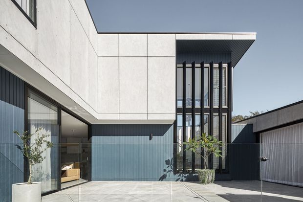Contemporary multi-level residential facade viewed from a paved courtyard. The building combines crisp white panelled upper walls, dark vertically ribbed cladding, extensive glazing, and a projecting flat roof. Tall narrow windows are screened by prominent black vertical fins, while a garage or ancillary volume sits to the right. The restrained modern composition is softened by a few potted plants and an outdoor seating area visible through the glazing.