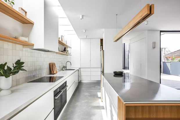 Bright contemporary galley kitchen with white flat-panel cabinetry, white tiled splashback, pale stone benchtops and a large island with timber detailing. A polished concrete floor, open timber shelving and a prominent exposed timber beam add warmth and texture. The kitchen opens toward an exterior driveway or courtyard through a large glazed opening.