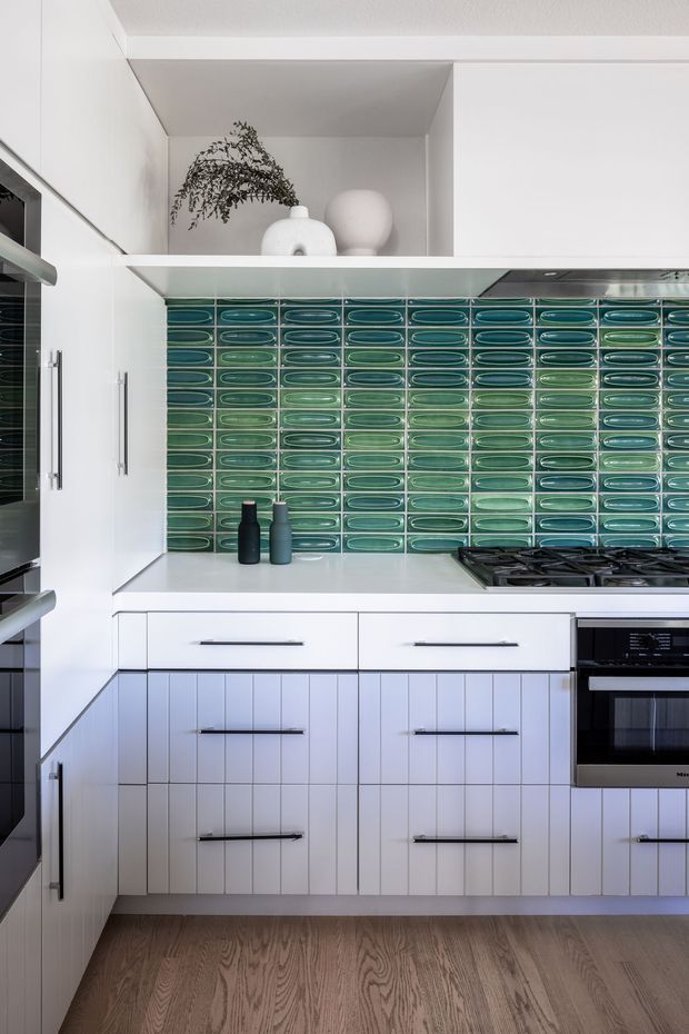 A compact contemporary kitchen with white vertically grooved cabinetry, long dark metal handles, a pale benchtop, and a striking glossy green-blue elongated tile splashback. Integrated appliances and a gas cooktop line the right side, while a small open shelf displays neutral ceramic vessels and greenery. The kitchen has warm timber flooring and clean, bright detailing.