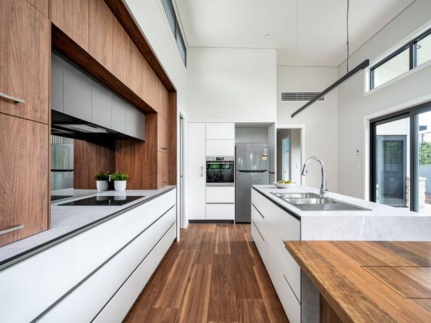 Bright contemporary kitchen with a long white island, integrated stainless-steel sink and tap, timber breakfast bar, extensive white cabinetry, and a warm timber feature wall with open shelving and built-in appliances. The room has a high ceiling, clerestory glazing, dark-framed windows and doors, and a polished, functional open-plan feel.