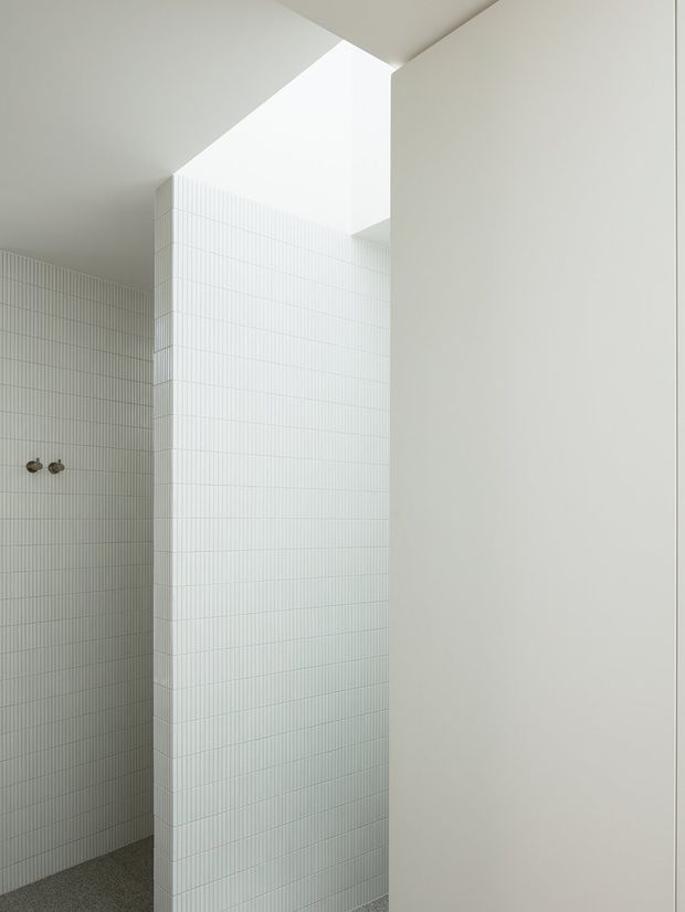 A minimalist bathroom interior featuring floor-to-ceiling white vertically lined ceramic wall tiles, a partially concealed shower area with wall-mounted fittings, smooth white plastered surfaces, and a high open ceiling or skylight that brings in diffuse natural light. The restrained palette and geometric forms create a clean, serene atmosphere.