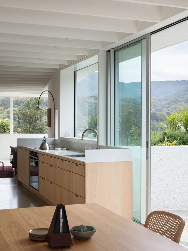 Bright, open-plan kitchen and dining area with extensive floor-to-ceiling glazing overlooking a lush mountainous landscape. The kitchen features light timber cabinetry, a pale stone benchtop and island, integrated appliances, a polished concrete floor, and exposed white ceiling beams. A timber dining table and woven chair occupy the foreground.