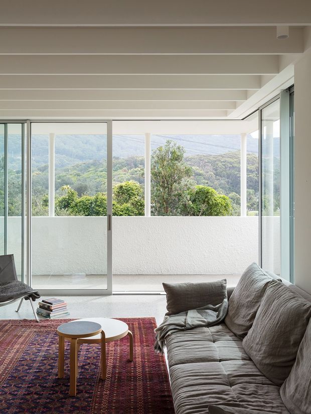 Bright contemporary living room with a large grey sofa, patterned area rug, nested round coffee tables, and a lounge chair. Full-height sliding glass doors open to a balcony or terrace with a white rendered balustrade and expansive green hillside views. The room has a white exposed-beam ceiling and abundant natural light.
