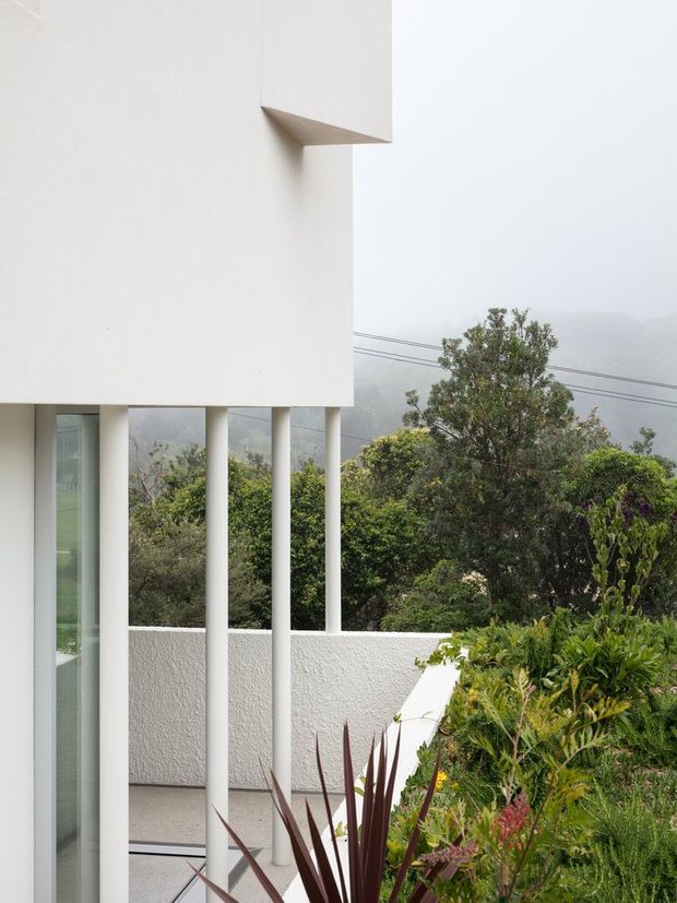 A minimalist white rendered building exterior with a projecting upper volume supported by slender rectangular columns. A glazed opening and terrace or balcony are visible, bordered by a low white parapet and dense landscaped planting. Misty hills and overhead utility lines appear in the background.