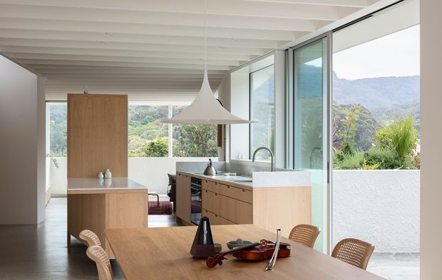 Bright open-plan kitchen and dining area with warm timber cabinetry, a central island, white benchtops, a large sculptural pendant light, and full-height glazing opening to a leafy mountainous landscape. The white exposed-beam ceiling and indoor-outdoor connection create an airy, contemporary feel.