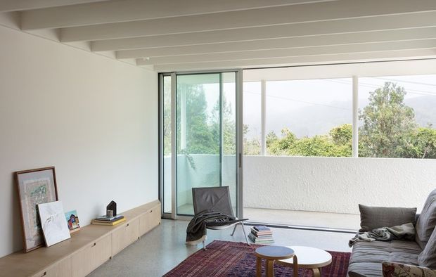 Bright modern living room with a low built-in timber media cabinet, sectional sofa, coffee tables, framed artwork, and a large sliding glass door opening to a covered outdoor area. The space features exposed white ceiling beams, polished concrete flooring, and expansive glazing with views of greenery beyond.