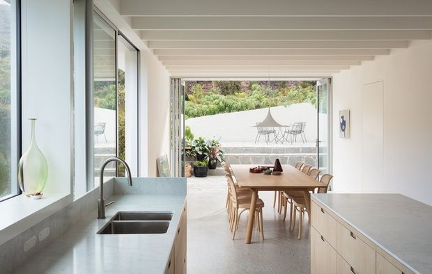 Bright open-plan kitchen and dining area with pale stone benchtops, light timber cabinetry, stainless steel sink and tapware, and a long timber dining table. Full-height glazing and an open connection lead to a landscaped outdoor terrace, while exposed white ceiling beams add architectural definition.