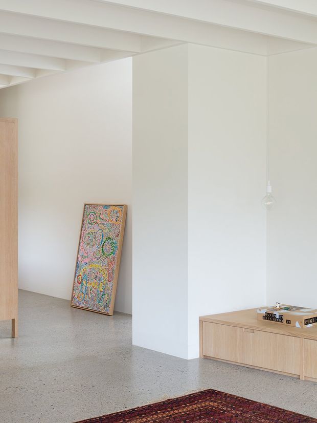 Bright minimalist interior with white walls, exposed white ceiling beams, pale timber built-in cabinetry and furniture, a polished light terrazzo or concrete floor, and a colourful framed artwork leaning against the wall. A small pendant light and patterned rug add subtle visual accents.