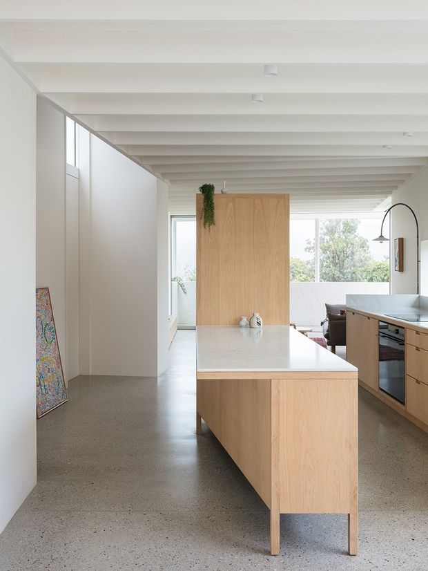 Bright open-plan kitchen with a central light timber island and pale stone benchtop, matching timber cabinetry, integrated appliances, polished concrete flooring, and a white exposed-beam ceiling. Large windows provide abundant natural light, with a glimpse of an adjoining living area.