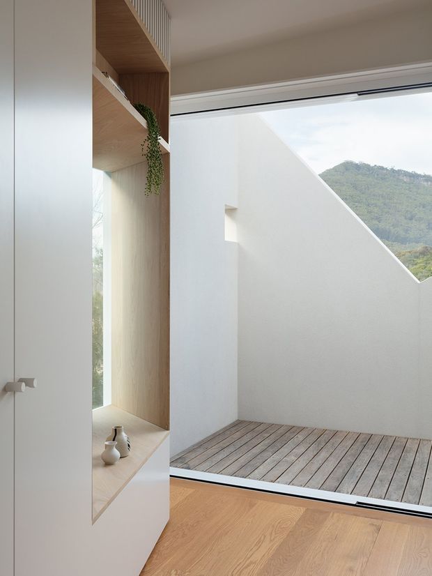A bright, minimalist interior corner with pale timber flooring and a built-in timber display niche beside expansive glazing. The glazing opens visually onto a small timber-decked courtyard or terrace enclosed by white walls, with a green hillside visible beyond. A trailing plant and a few small ceramic objects add subtle decoration.