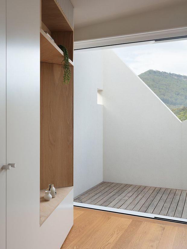 A minimalist interior adjoining a small private courtyard, featuring pale timber flooring, a built-in white and light-wood shelving unit, and a large glazed opening. The courtyard has a timber deck enclosed by crisp white walls with a diagonal opening framing a green hillside beyond.