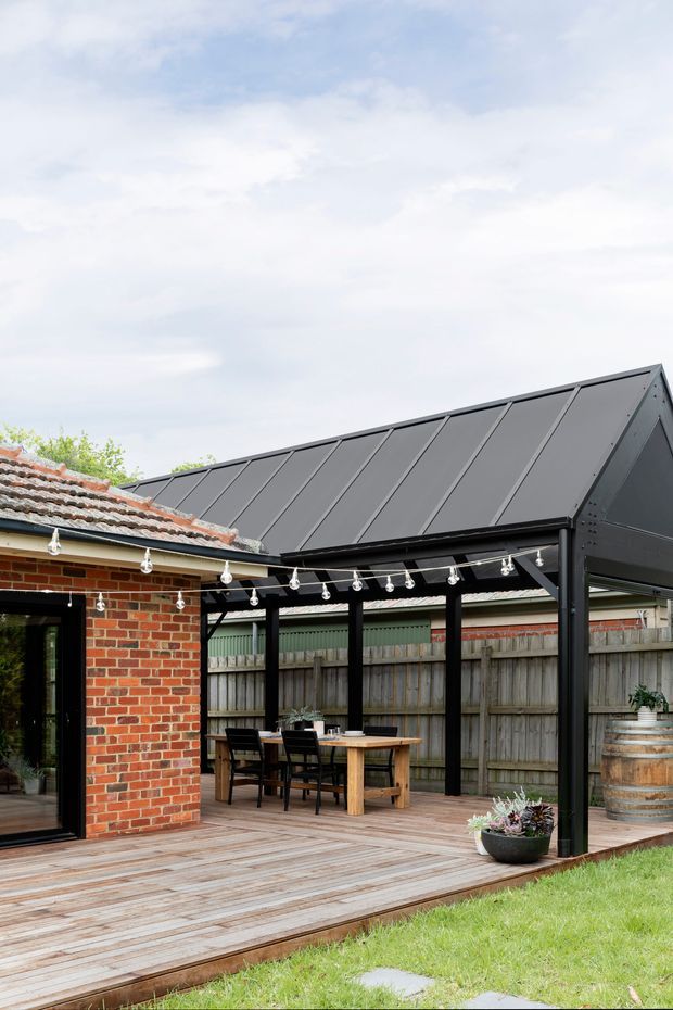A covered outdoor entertaining area adjoining a red brick building, with a dark steel-framed gable roof, timber decking, dining table and chairs, festoon lights, timber fencing, and garden elements. The image focuses on the outdoor patio and pergola structure.
