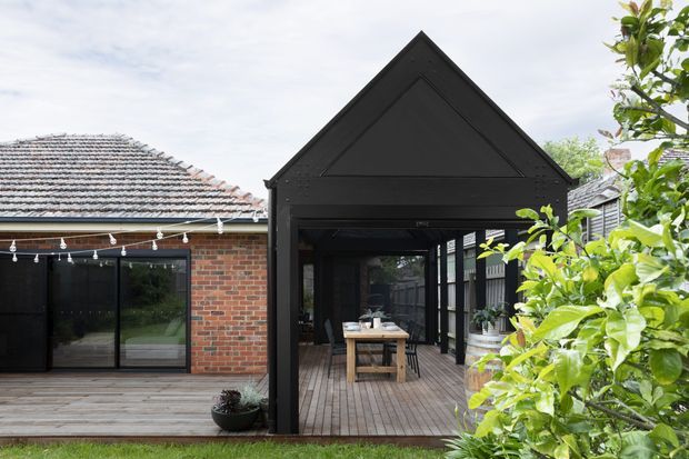 Rear exterior view of a renovated brick home with a prominent black, gabled outdoor pavilion or extension opening onto a timber deck. The covered space contains a dining table and chairs, with large glazed doors connecting to the house and a leafy garden surrounding the deck.