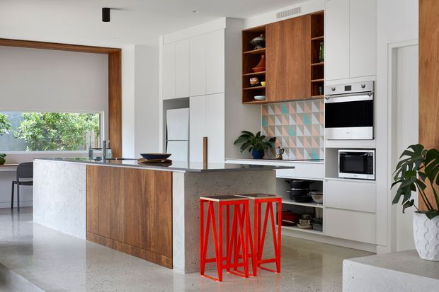Bright contemporary kitchen with a large island featuring a terrazzo or stone benchtop and warm timber cabinetry. White full-height joinery integrates the refrigerator and ovens, while open timber shelving displays kitchenware. A colourful geometric tile splashback adds a patterned feature, and vivid red bar stools provide contrast. The kitchen opens to a dining or living area and includes indoor plants.