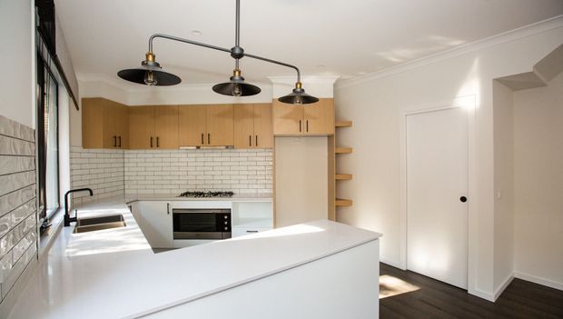 Bright contemporary kitchen with a large white island and integrated sink, timber cabinetry, white subway-tile splashback, stainless-steel appliances, dark timber flooring, and an industrial-style three-light pendant. A hallway or entry area is visible to the right.