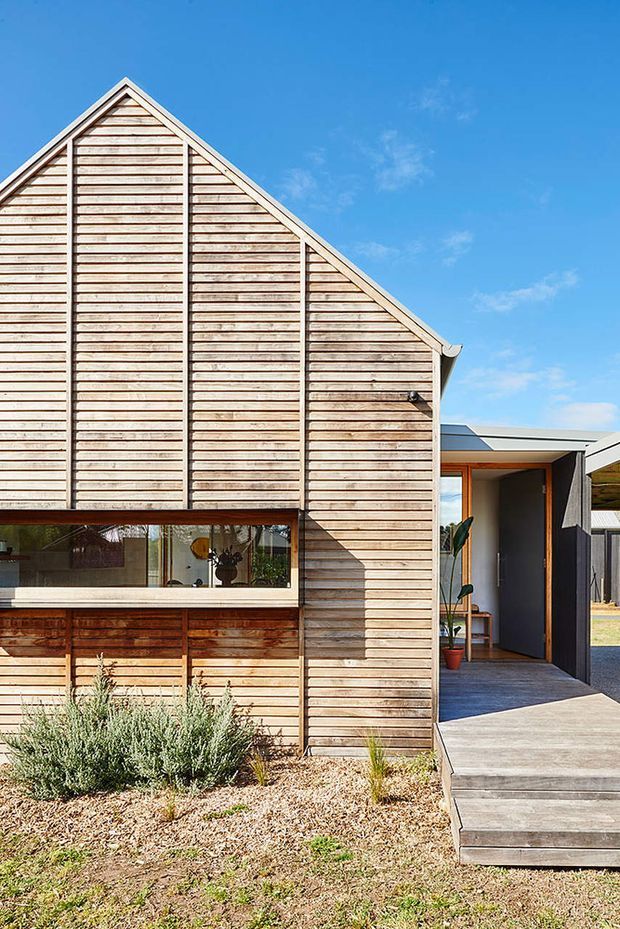 Exterior view of a contemporary gabled building clad in horizontal natural timber weatherboards, with a prominent triangular gable, vertical structural battens, large glazed window, and a recessed entry porch. The home is set beside a simple gravel and planted landscape, with timber steps leading to the entrance and a connected flat-roofed volume visible behind.
