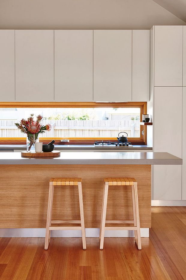 Bright, compact contemporary kitchen with a timber island and two simple timber bar stools. The kitchen features extensive matte off-white overhead and full-height cabinetry, a pale stone-look benchtop, warm timber flooring, and a long timber-framed horizontal window above the cooktop and rear counter. Natural light fills the space, with restrained styling and a small floral arrangement on the island.