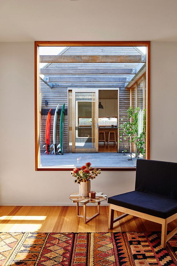A bright, compact living area with warm timber flooring, a large patterned geometric rug, a black upholstered timber-framed chair, and a small round glass-topped side table with flowers. A large timber-framed internal window looks onto a landscaped courtyard or outdoor deck, with surfboards, horizontal timber cladding, exposed beams, and glazed double doors visible beyond.