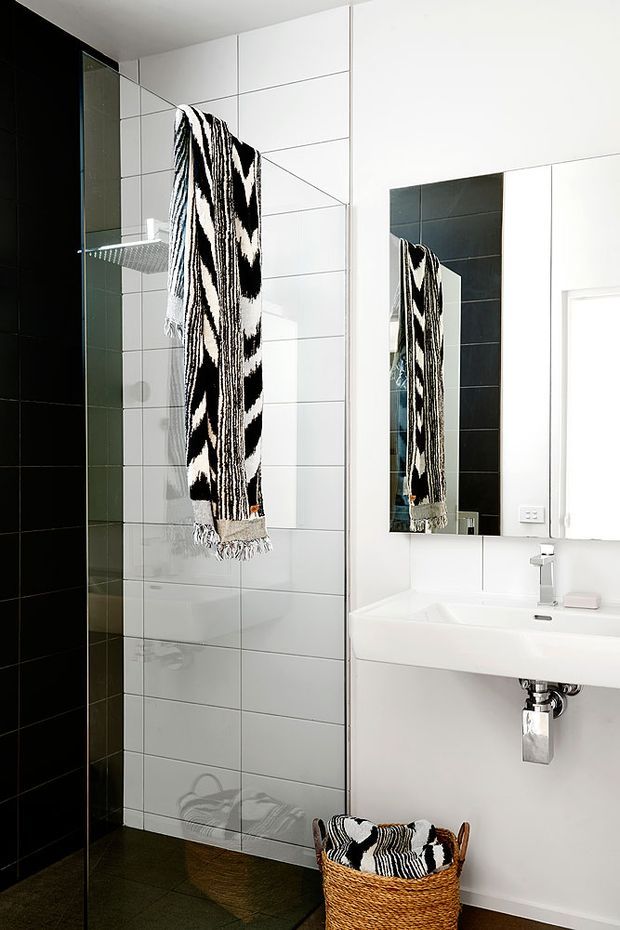 Bright contemporary bathroom with a frameless glass shower enclosure, large-format white wall tiles, a black tiled shower feature wall, wall-mounted white basin, large rectangular mirror, and chrome tapware. A bold black-and-white patterned towel hangs over the shower glass, with a woven basket below.