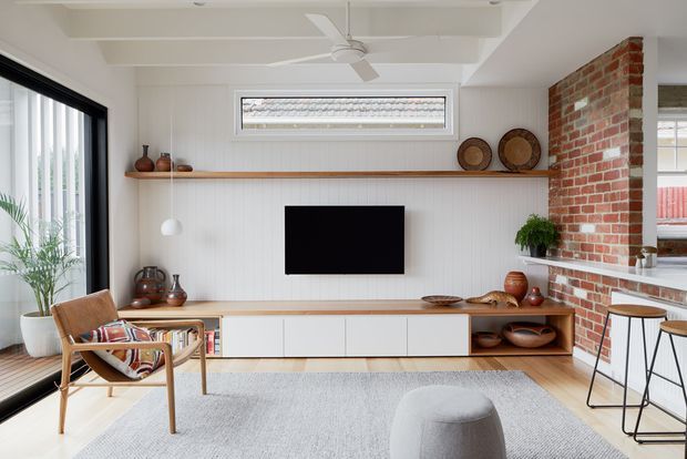Bright open-plan living room with a white paneled media wall, long timber floating shelf and low white built-in TV cabinetry. A red brick feature wall separates the living area from a kitchen or bar zone, while large glazing, a ceiling fan, woven timber seating, indoor plants and neutral furnishings create a relaxed contemporary interior.