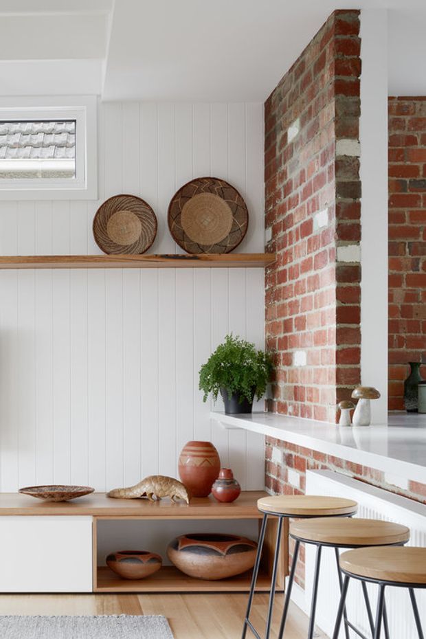 A bright, warm kitchen interior featuring white vertical wall panelling, exposed red brickwork, a long timber display shelf, low timber cabinetry, and a white stone benchtop with a raised brick partition. Three timber-topped stools line the breakfast counter. Woven wall baskets, ceramic vessels, a potted plant, and sculptural decor add a natural, relaxed character. A small window with a tiled roof view provides daylight.
