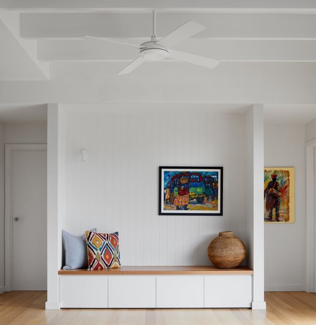 Bright, minimalist interior featuring a built-in white wall unit with vertical paneling, a timber bench seat, and low white cabinetry. Two cushions, woven decorative vessels, and colourful framed artwork add warmth and personality. Pale timber flooring, white walls, a modern ceiling fan, and doorways to adjacent rooms create a clean, airy residential setting.