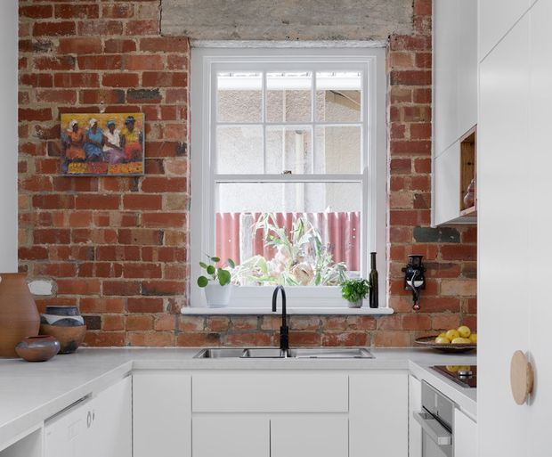 A compact, bright kitchen with exposed red brick walls, white cabinetry, pale benchtops, and a central multi-pane window above a stainless-steel sink. A matte black gooseneck tap, open shelving, small plants, and understated ceramic objects add contrast and warmth. The room combines original masonry character with clean contemporary joinery.