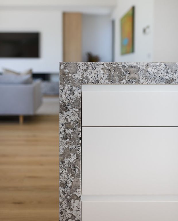 Close-up architectural interior detail showing a white kitchen island or built-in cabinetry with a heavily patterned grey-and-white natural stone benchtop and waterfall edge. The open-plan living room beyond includes light timber flooring, a pale sofa, wall-mounted television, and white walls. The image focuses on the stone edge and crisp white joinery.