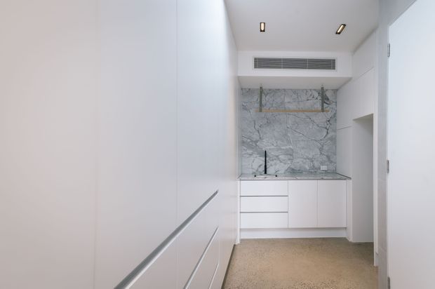 A compact, bright minimalist kitchen or kitchenette with extensive white handleless cabinetry, a pale stone benchtop, and a dramatic grey-veined stone splashback extending up the rear wall. Open glass shelving, integrated ventilation, recessed ceiling lights, and a warm neutral floor complete the streamlined fit-out.