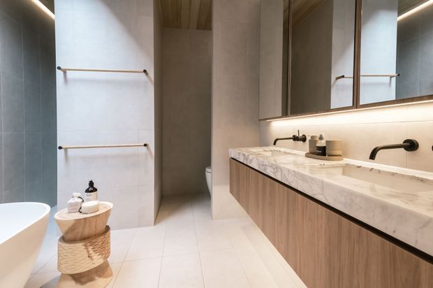 Contemporary bathroom with a freestanding white bathtub, walk-in shower, double vanity, large illuminated mirror cabinet, pale stone surfaces, warm timber cabinetry, and brushed dark metal fixtures. The design is clean, warm, and refined with concealed linear lighting.